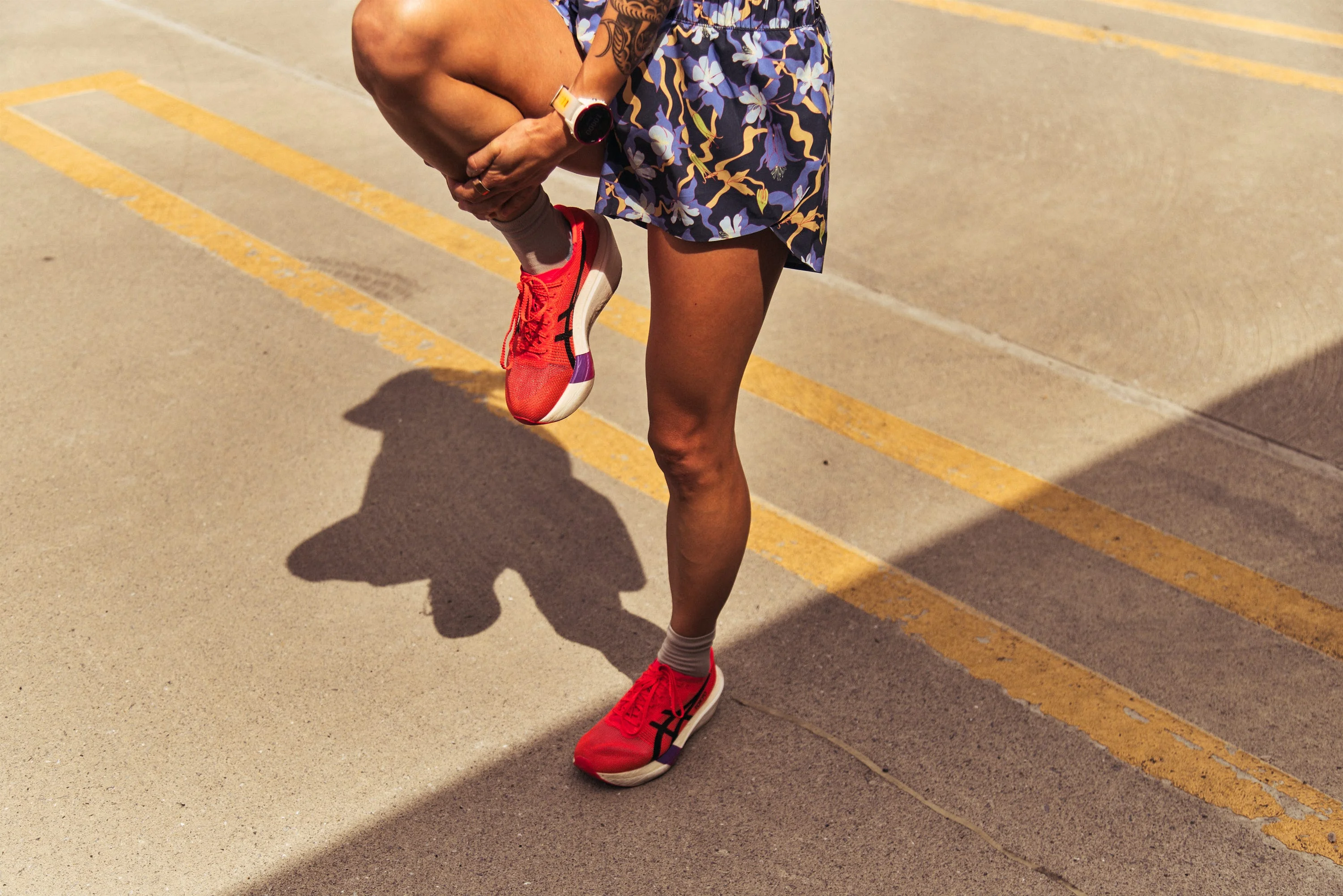 Person in floral shorts stretching leg and wearing red running shoes.