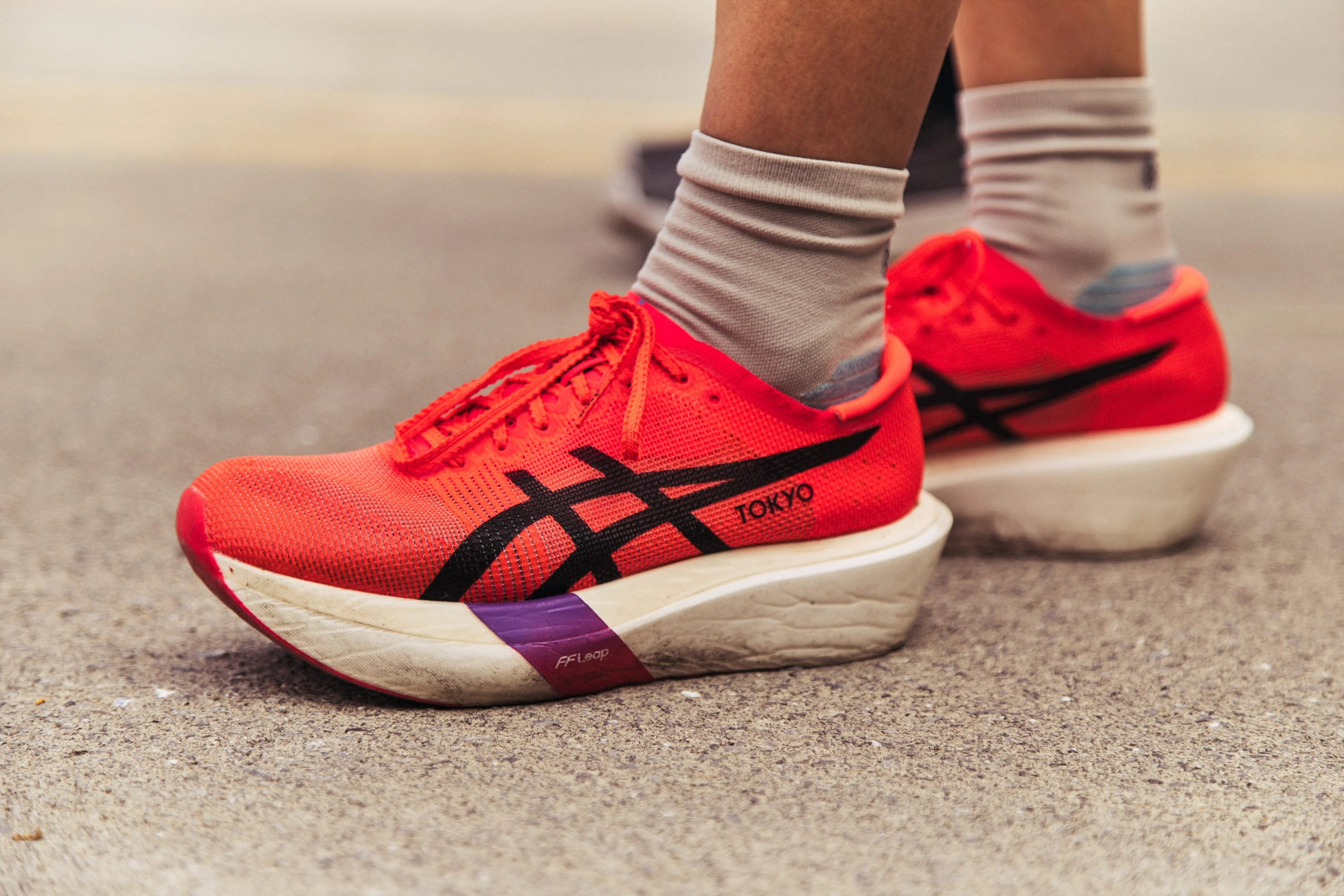 Bright orange running shoes with thick white soles worn by a person with gray socks on pavement.