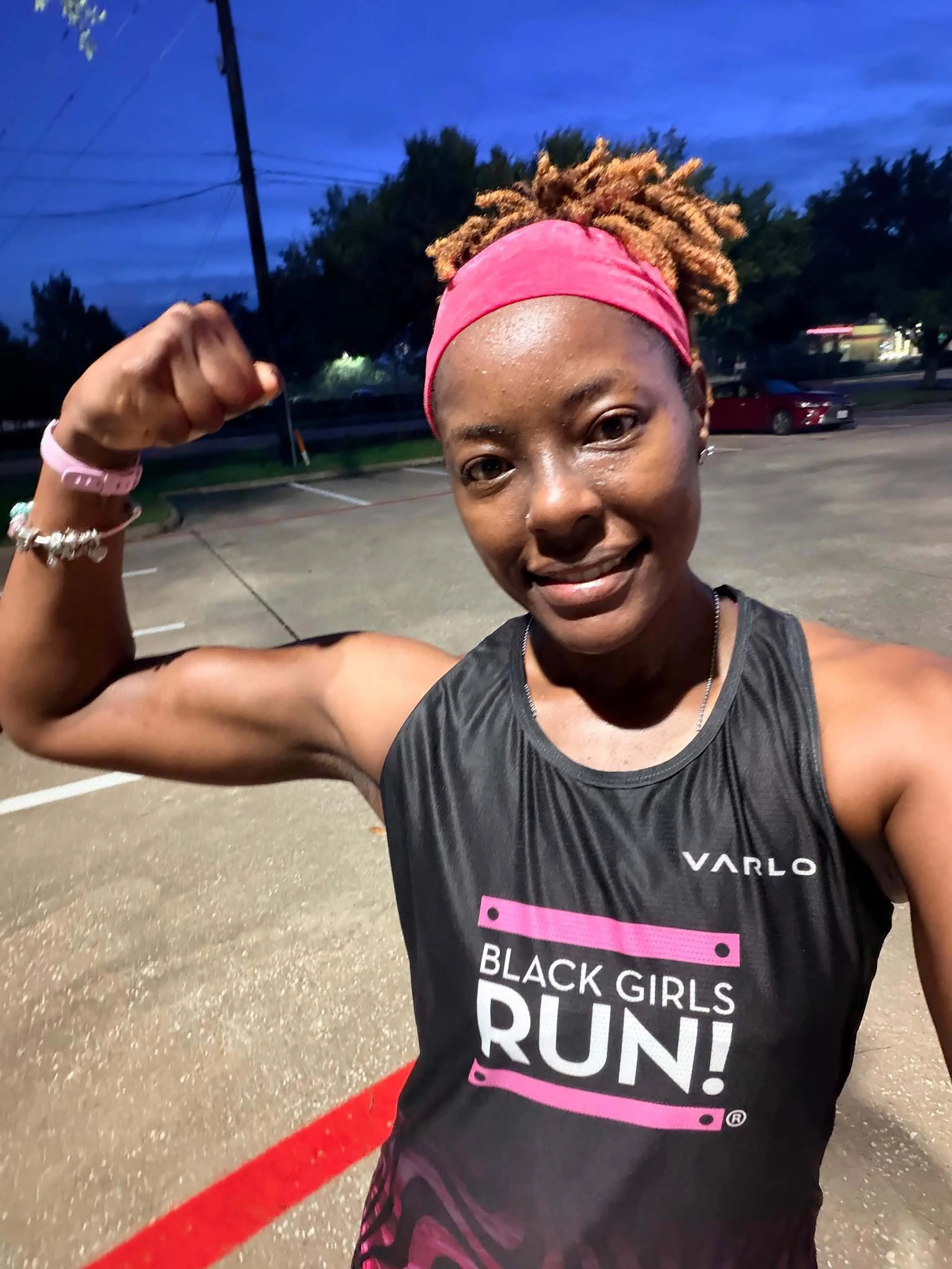 Runner smiling, wearing "Black Girls RUN!" tank top, flexing arm in outdoor setting.