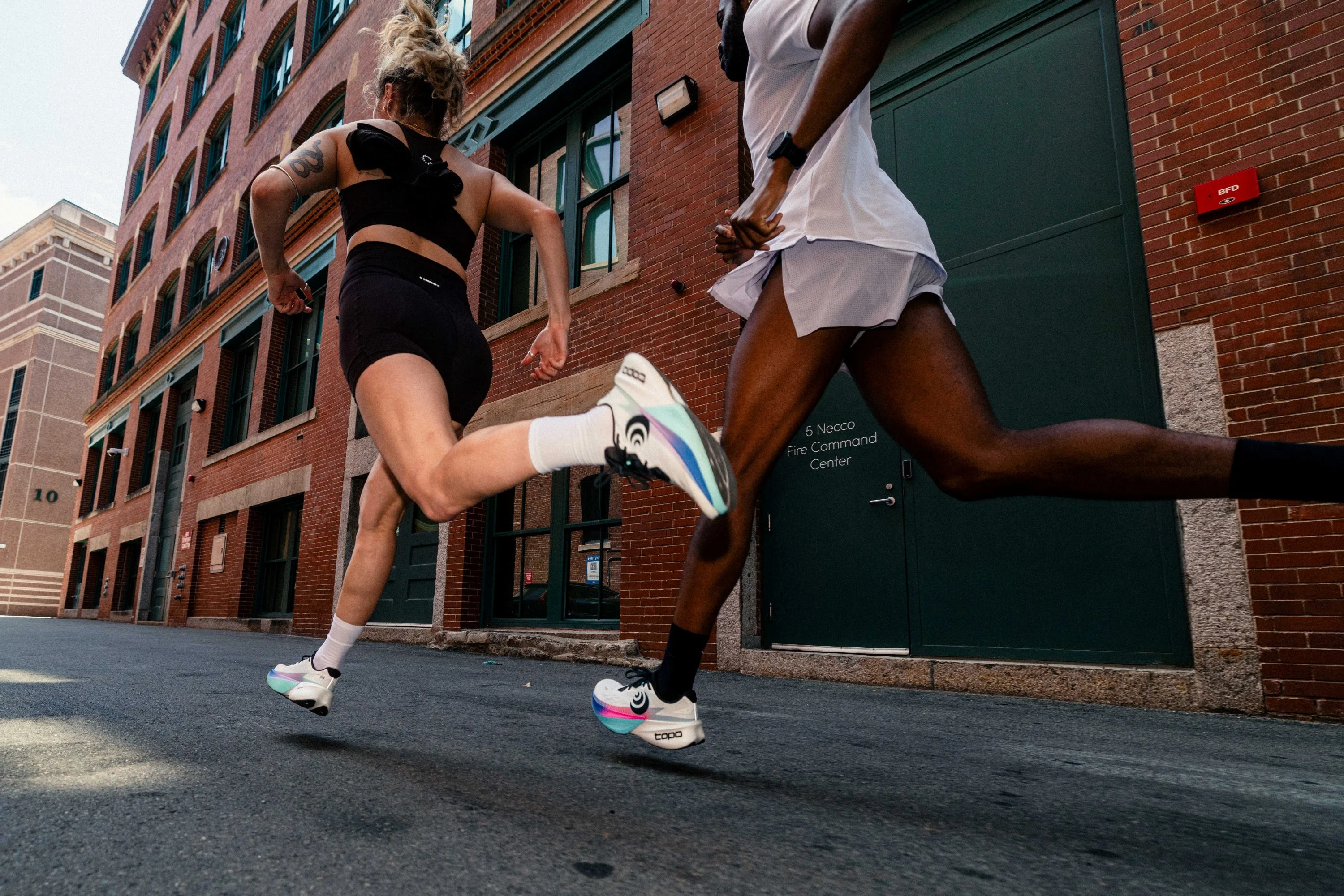 Two people running past brick buildings in athletic gear. Two people running past brick buildings in athletic gear.