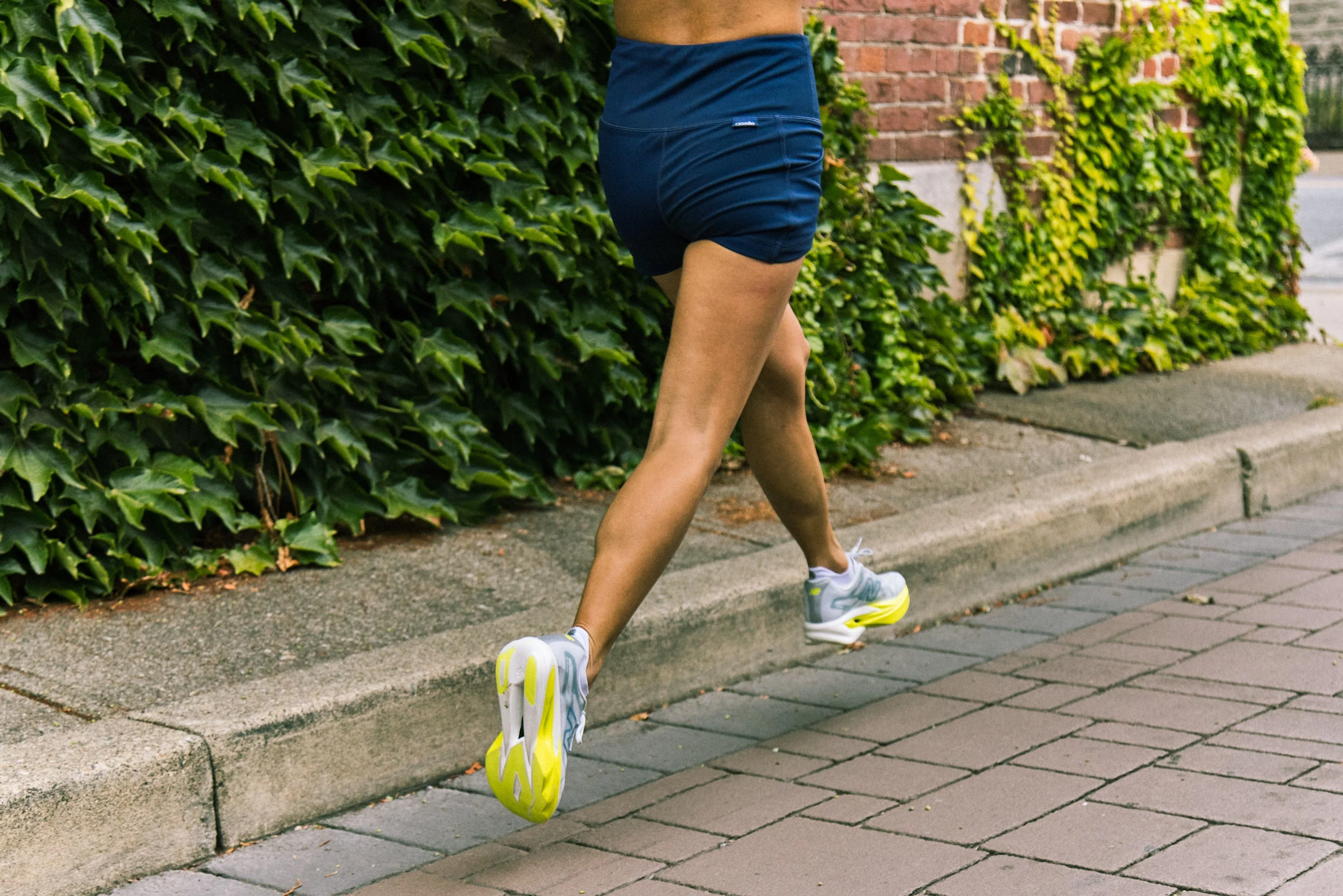 Person jogging along a sidewalk beside a wall covered in ivy, wearing athletic shorts and shoes. Person jogging along a sidewalk beside a wall covered in ivy, wearing athletic shorts and shoes.