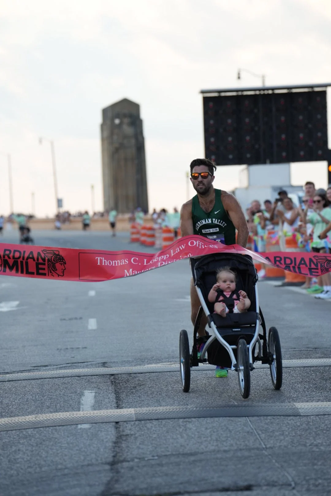 Runner pushing a stroller with a child crosses the finish line at a race, with spectators cheering in the background.