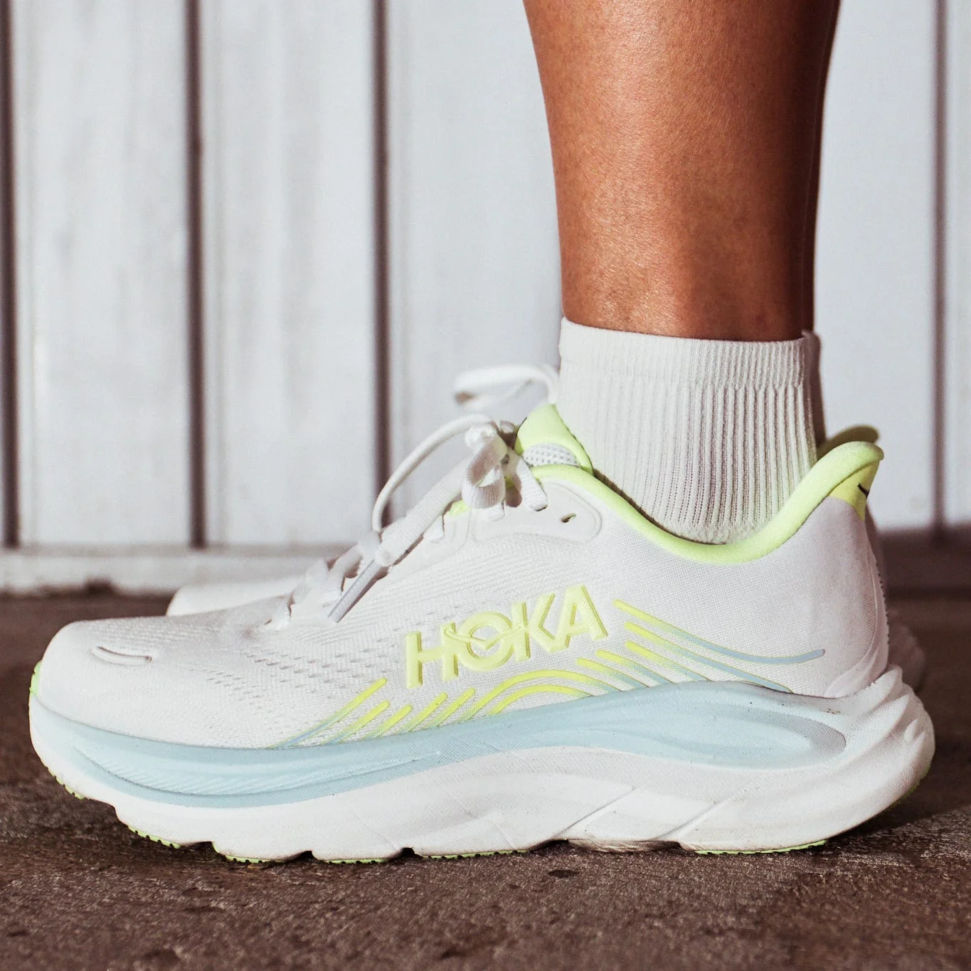 HOKA running shoes with a white and neon green design, worn on a person's feet against a textured surface. HOKA running shoes with a white and neon green design, worn on a person's feet against a textured surface.