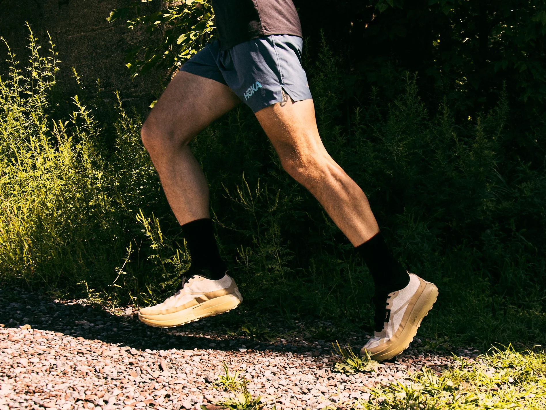 Runner in athletic gear wearing Norda 055 shoes jogging on a gravel path surrounded by green foliage.