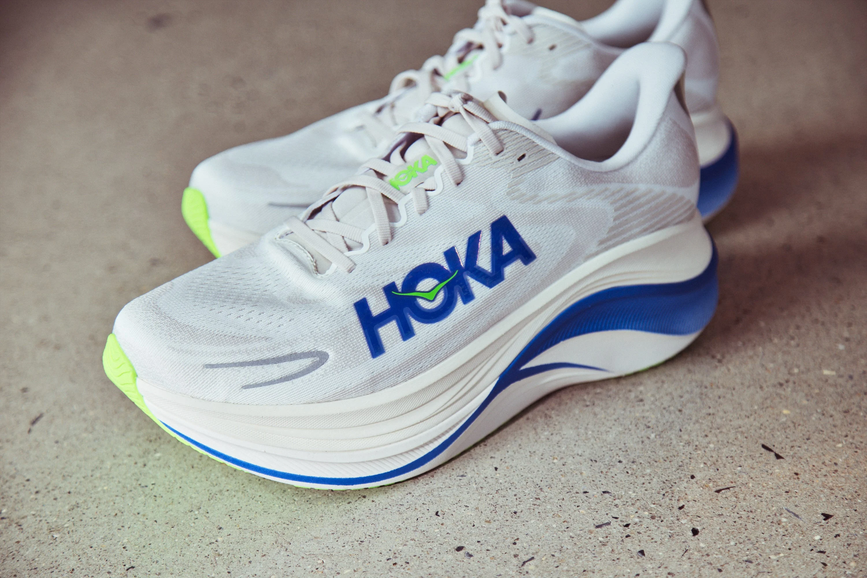 Hoka running shoes in white with blue accents and neon green soles, featuring a sleek design and prominent branding. Hoka running shoes in white with blue accents and neon green soles, featuring a sleek design and prominent branding.