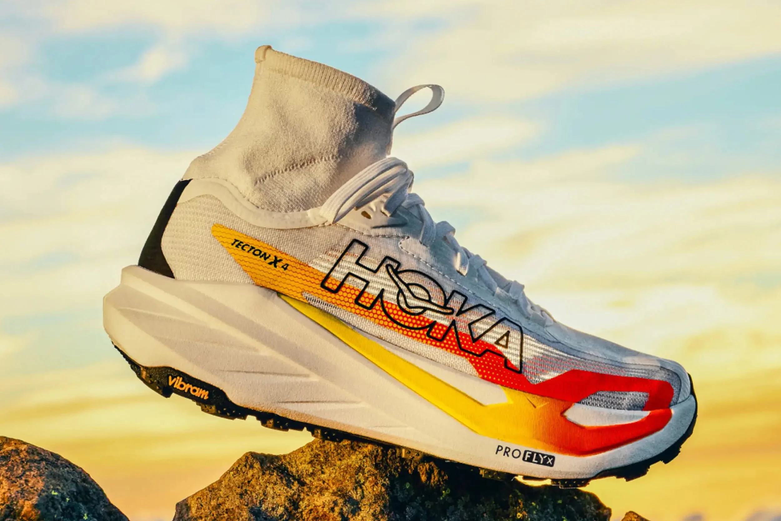 Dynamic Hoka Tecton X 4 running shoe in white with orange and yellow accents on a rocky surface against a colorful sky. Dynamic Hoka Tecton X 4 running shoe in white with orange and yellow accents on a rocky surface against a colorful sky.