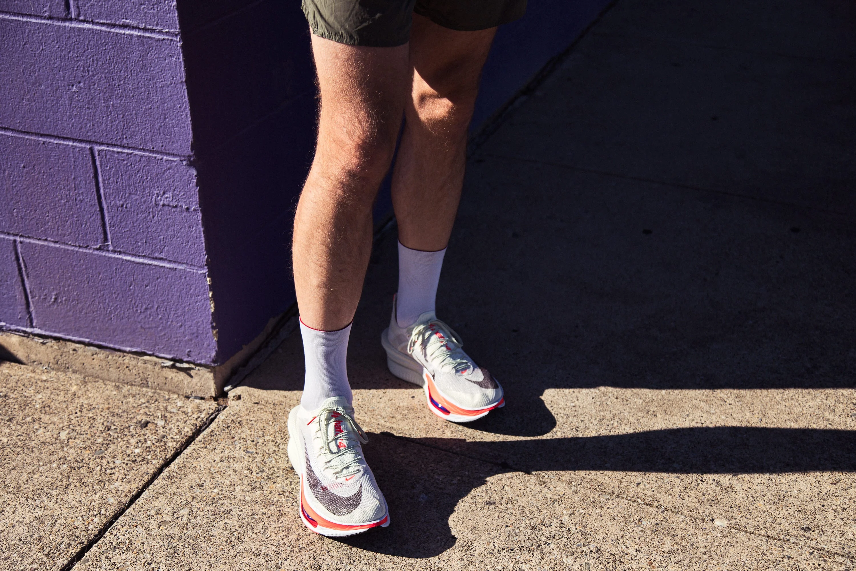 A person wearing athletic shoes and white socks standing on pavement near a purple wall.