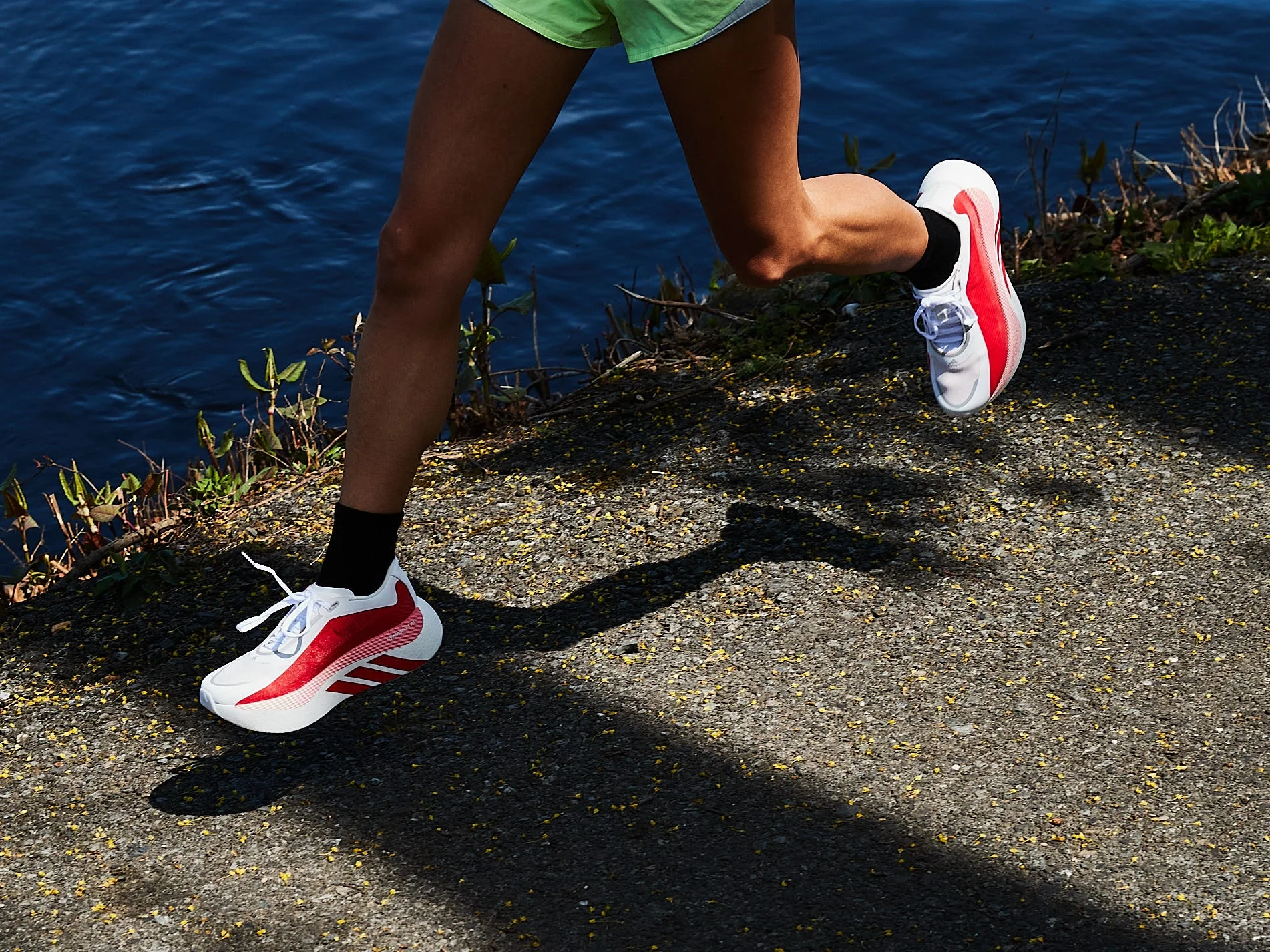 A runner in red and white shoes jogs along a path near water, with shadows cast on the ground.