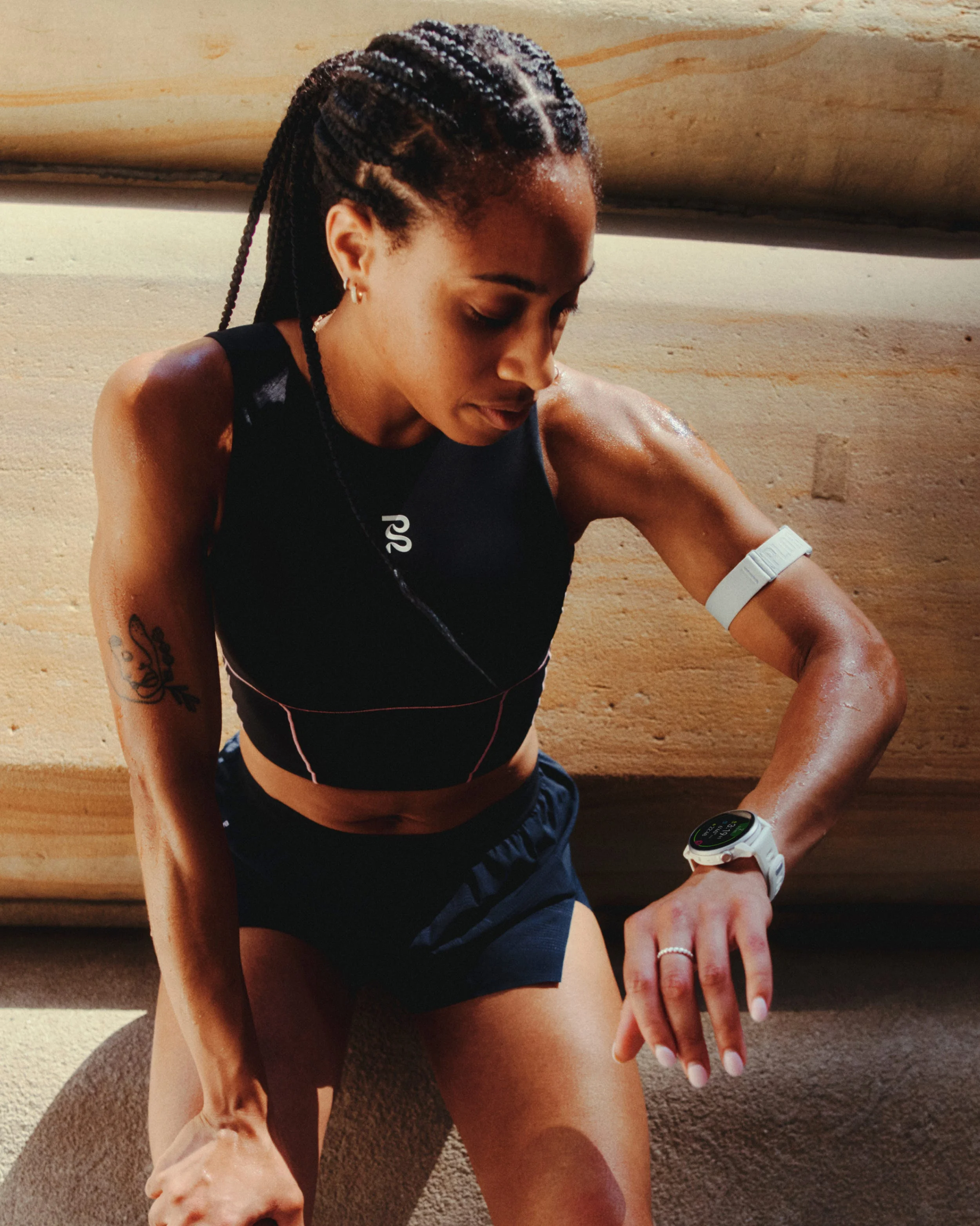 Athlete checking fitness watch while seated, wearing sports attire, with braided hair and tattoos.