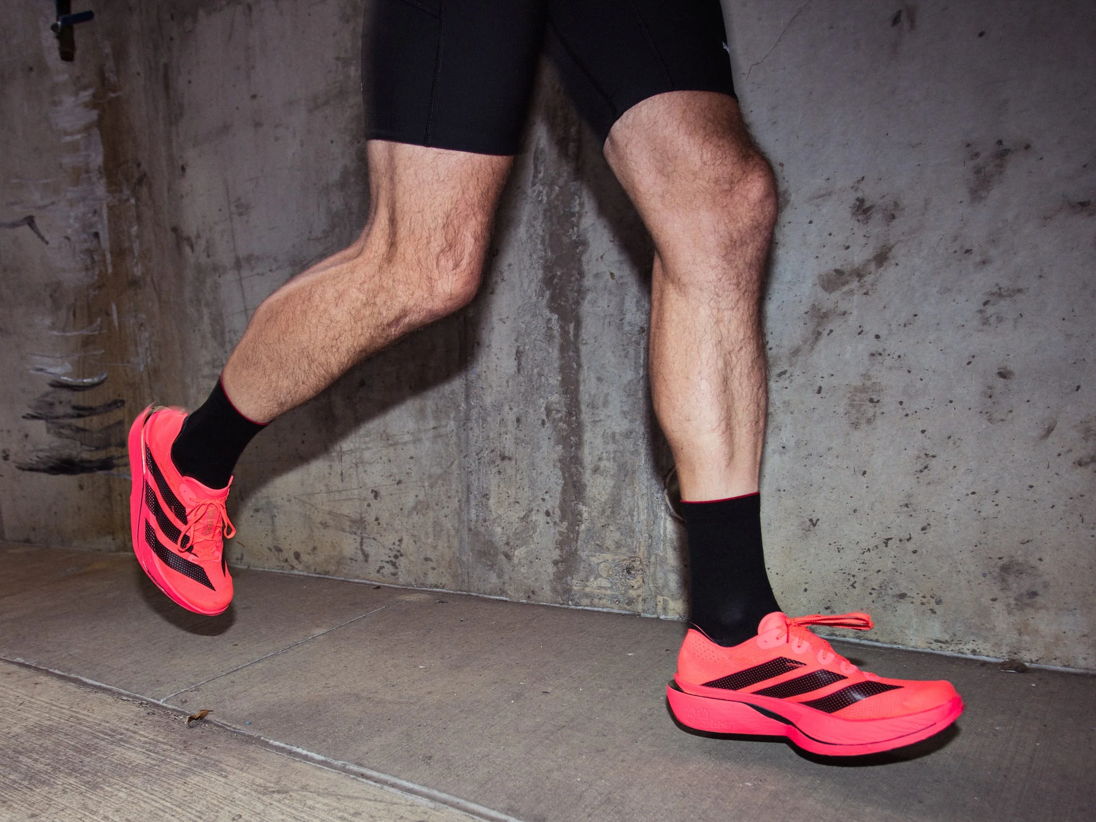 Runner wearing bright pink Adizero Adios Pro 5 shoes and black shorts, jogging on a concrete surface.