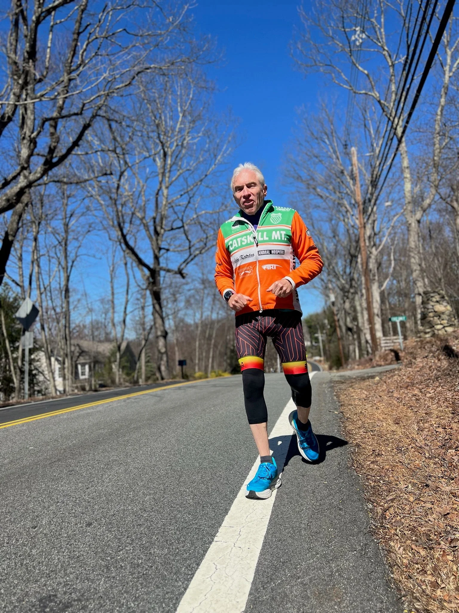 Man running on a sunny road wearing an orange and green jacket, colorful shorts, and bright blue shoes.
